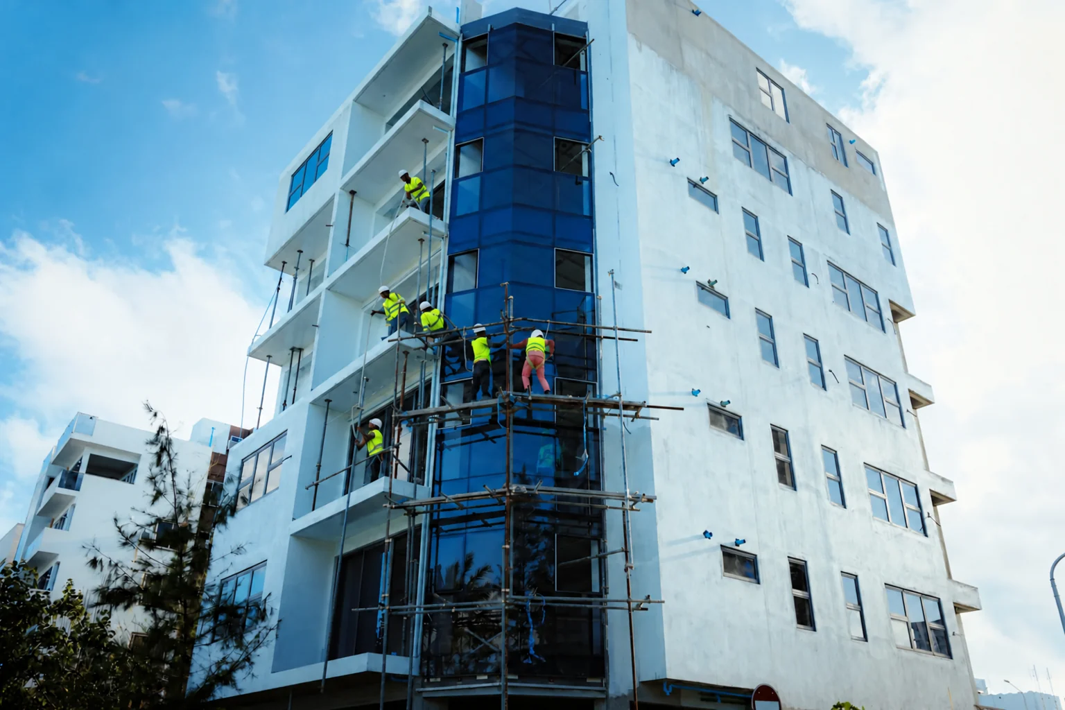 Construction workers in yellow safety vests and white helmets installing a modern glass facade.